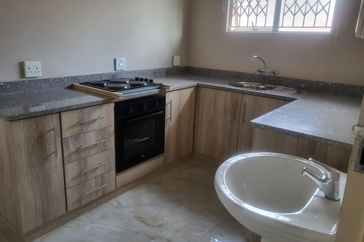 A functional kitchen featuring light wood-grain cabinetry, stone-look countertops, a built-in oven, electric hob, and a stainless steel sink with tiled flooring.