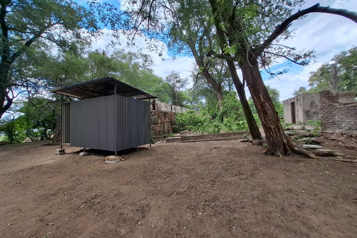 A view of a rustic outdoor area featuring a corrugated metal structure, stone boundary walls, and mature trees on an undeveloped plot.