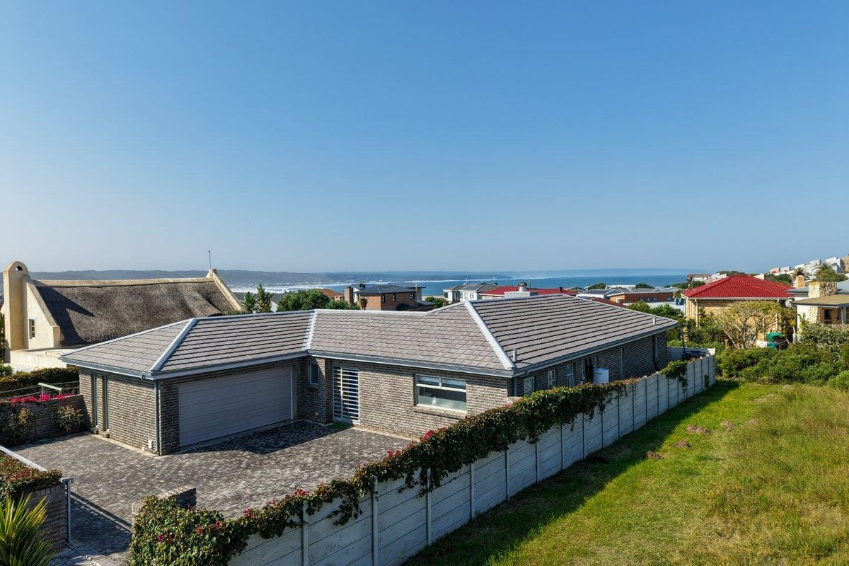 Aerial view of a single-story brick house featuring a tiled roof, paved driveway, double garage, and perimeter boundary wall with ocean views in the distance.
