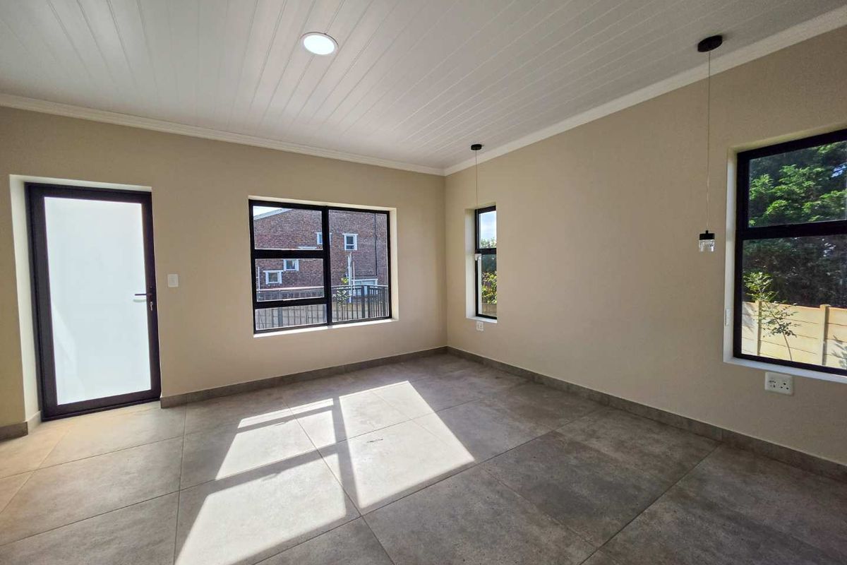 A bright, empty living area featuring large format floor tiles, a modern tongue and groove ceiling, and dark aluminium window frames.