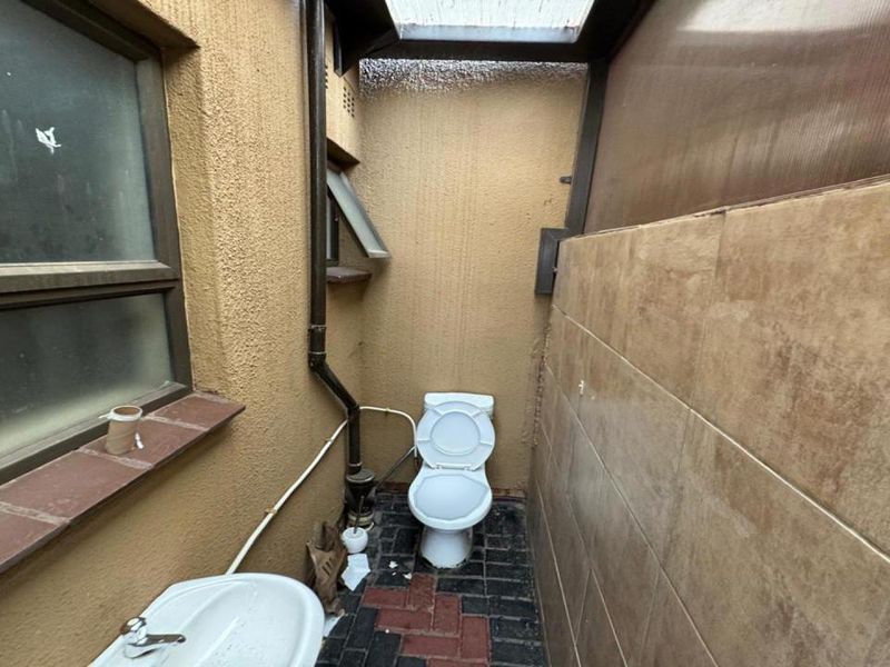 A small, outdoor-style bathroom featuring a toilet, pedestal basin, tiled walls, and a translucent roof panel.