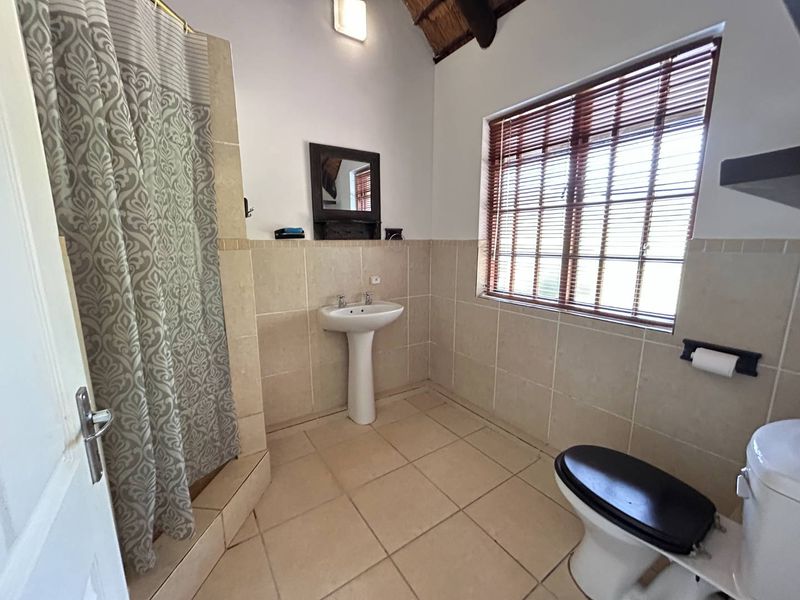 A functional bathroom featuring a pedestal basin, shower, toilet, tiled finishes, and a characterful exposed thatch ceiling with natural light.