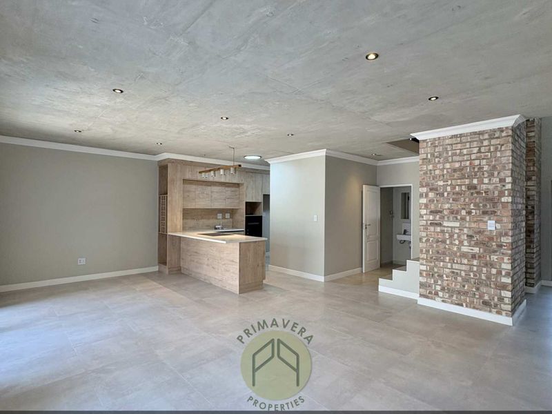 Spacious open-plan living area featuring an exposed concrete ceiling, modern kitchen cabinetry, and a striking exposed brick feature wall with neutral tiled flooring.