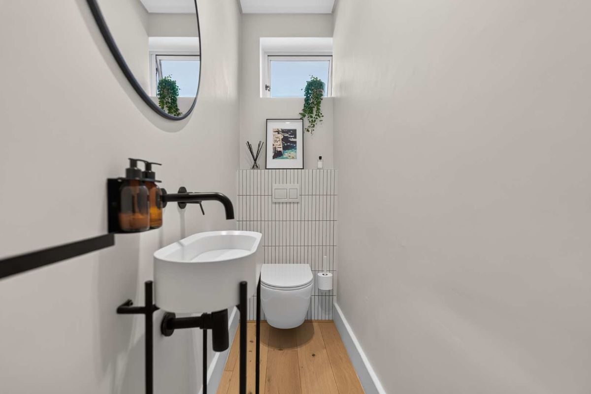 A modern, compact powder room featuring a wall-mounted toilet, vertical tile detailing, timber flooring, and a sleek pedestal sink with matte black fixtures.