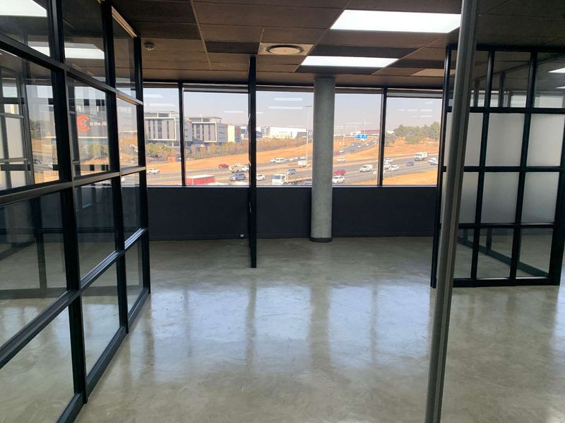 Modern open-plan office space featuring polished concrete floors, black metal-framed glass partitions, and expansive windows with highway views.
