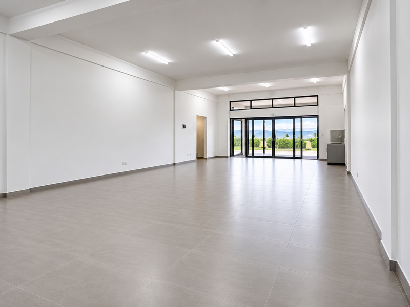 A spacious, modern open-plan office area featuring neutral tiled flooring, recessed lighting, and large sliding glass doors leading to an outdoor view.
