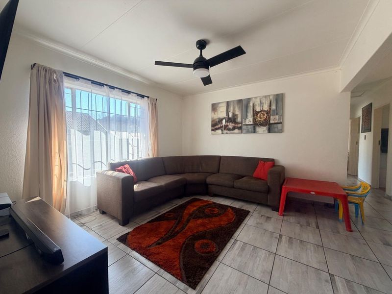 A living room featuring tiled flooring, a ceiling fan, and a large window with security bars, providing a functional and well-lit living space.