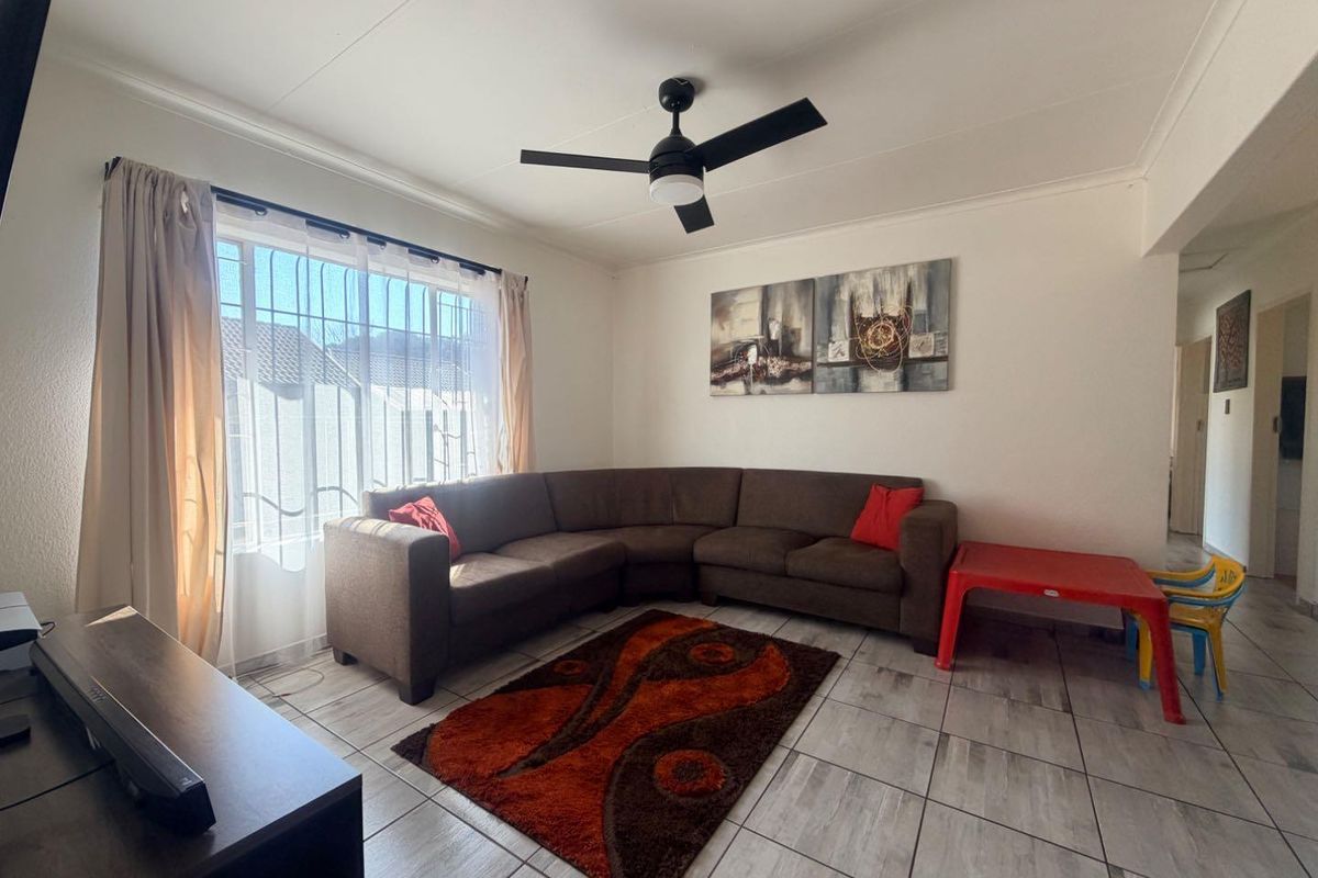 A living room featuring tiled flooring, a ceiling fan, and a large window with security bars, providing a functional and well-lit living space.