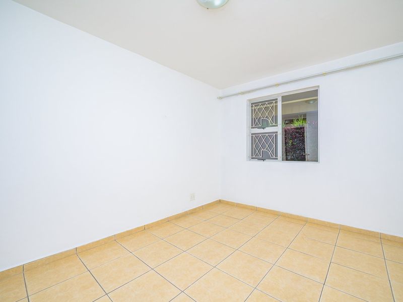A clean, empty bedroom featuring neutral tiled flooring, white walls, and a window fitted with security bars and a curtain rail.
