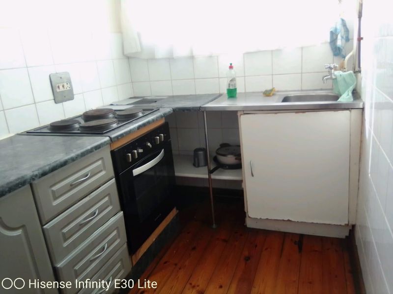 A compact kitchen featuring a built-in oven, electric hob, stainless steel sink, and tiled backsplash, requiring cosmetic updates to the cabinetry.