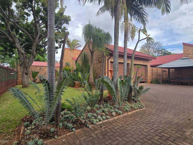 Front exterior view featuring a brick facade, double garage, paved driveway, and established garden with mature trees and palms.