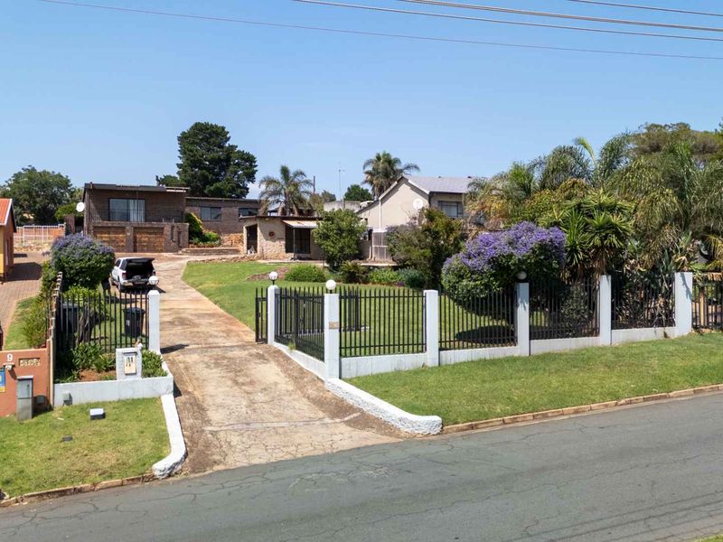 Front view of the property featuring a concrete driveway, established garden, and secure metal perimeter fencing with masonry pillars.