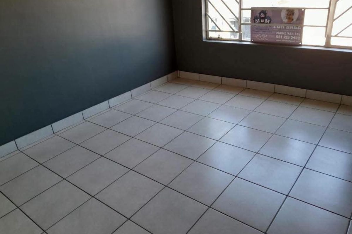 An empty living area featuring neutral tiled flooring and a window fitted with security bars.