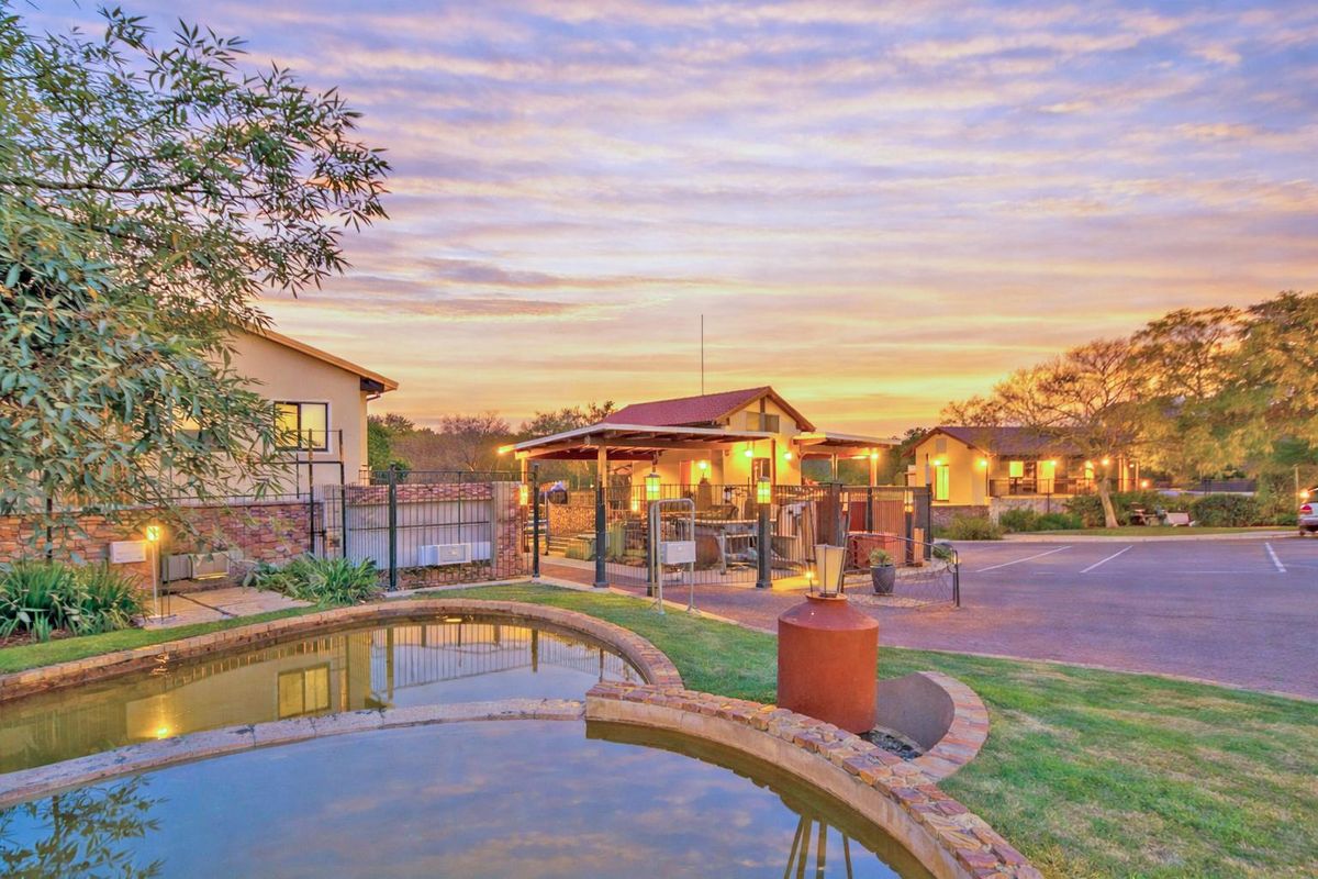 Exterior view of the apartment complex grounds featuring a landscaped pond, paved parking area, and covered outdoor communal facilities at sunset.