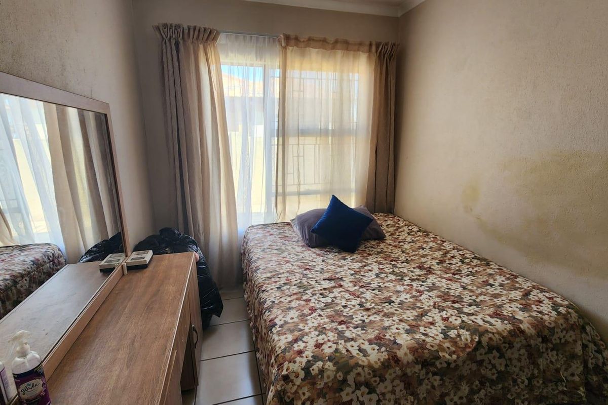 A compact bedroom featuring tiled flooring and a window with curtain treatments, showing signs of wall surface damage.
