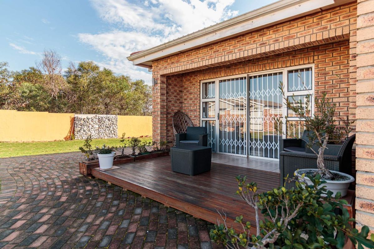 A rear exterior view featuring a wooden deck, brick facade, and sliding glass doors with security gates leading to a landscaped garden area.