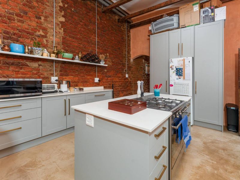 A modern industrial-style kitchen featuring exposed brick walls, grey cabinetry, a central island with a gas hob, and polished concrete flooring.