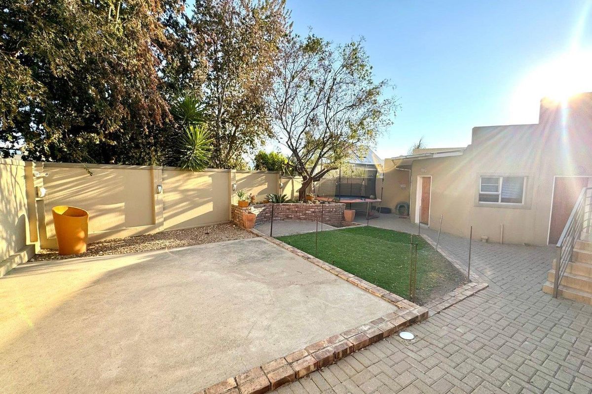 A rear exterior view featuring a paved patio area, a small artificial lawn, and a perimeter wall with established trees.