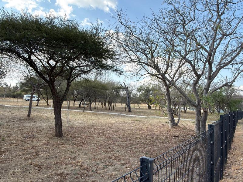 A view of a vacant commercial land plot featuring mature trees and secure wire mesh perimeter fencing.