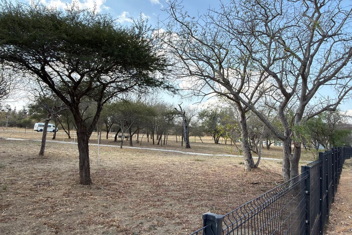 A view of a vacant commercial land plot featuring mature trees and secure wire mesh perimeter fencing.