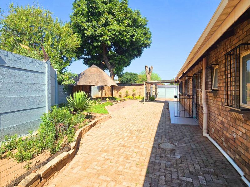 Rear exterior view featuring a paved courtyard, facebrick walling, security bars on windows, and a thatch lapa in the garden area.