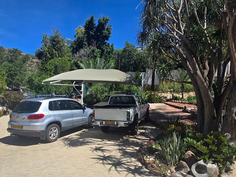 A paved front driveway featuring a shade sail carport, mature trees, and established garden landscaping.