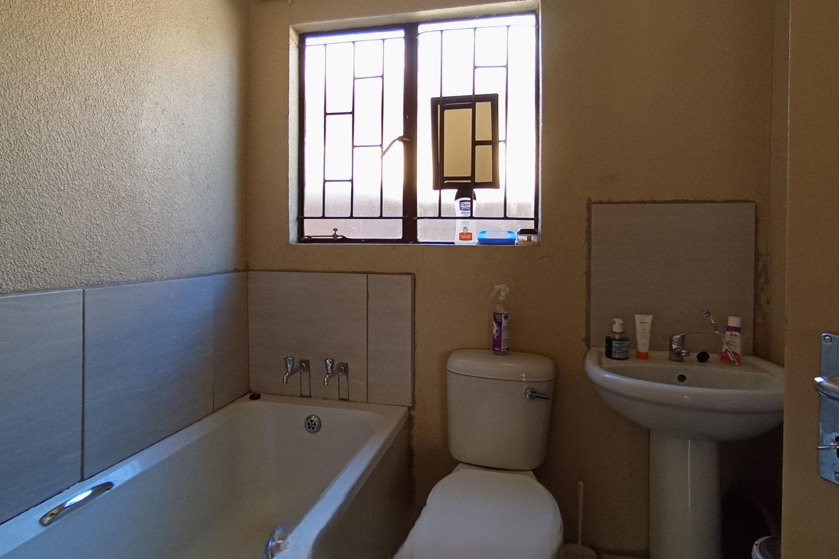 A functional bathroom featuring a standard bathtub, pedestal basin, and security-barred window, providing essential amenities with a dated aesthetic.