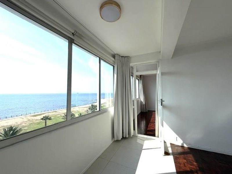 A bright, enclosed balcony featuring large windows with expansive ocean views, tiled and parquet flooring, and direct access to the interior living space.