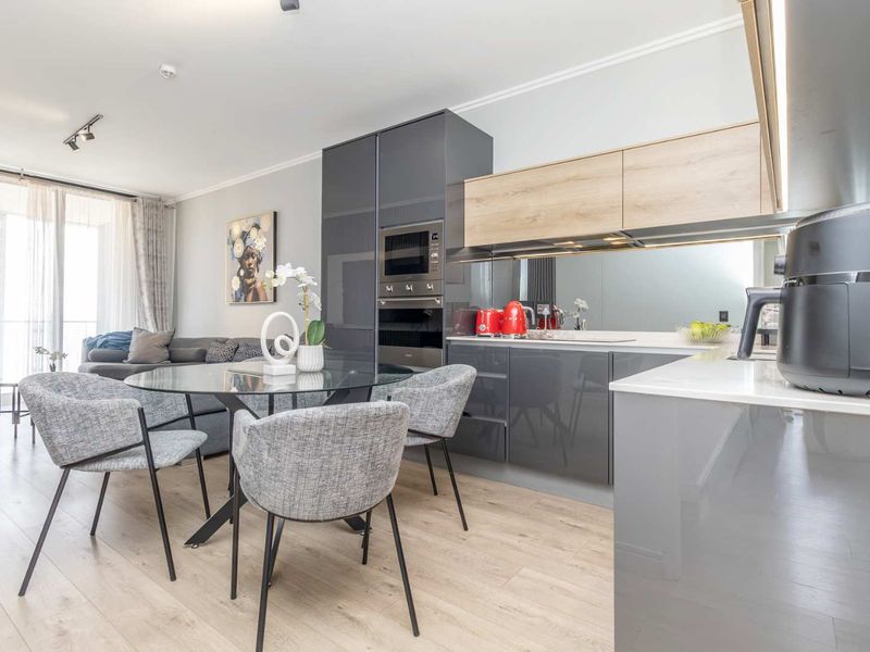 Modern open-plan living area featuring a sleek kitchen with integrated appliances, stone countertops, mirrored splashback, and light wood-look flooring leading to a balcony.