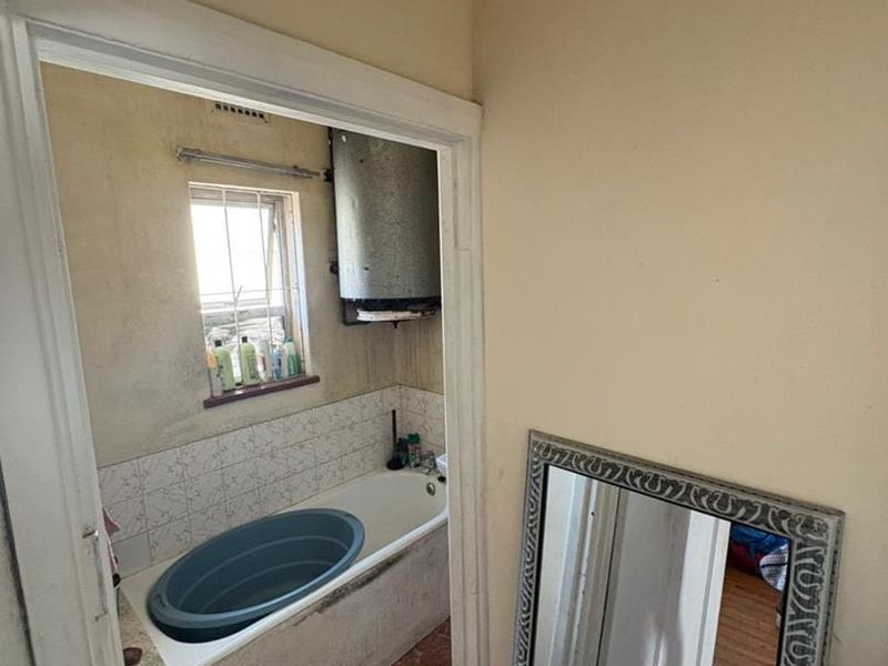 A dated bathroom featuring a built-in bathtub, tiled splashback, and wall-mounted geyser, requiring significant renovation and cosmetic updates to improve its condition.