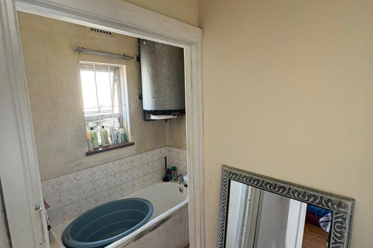 A dated bathroom featuring a built-in bathtub, tiled splashback, and wall-mounted geyser, requiring significant renovation and cosmetic updates to improve its condition.