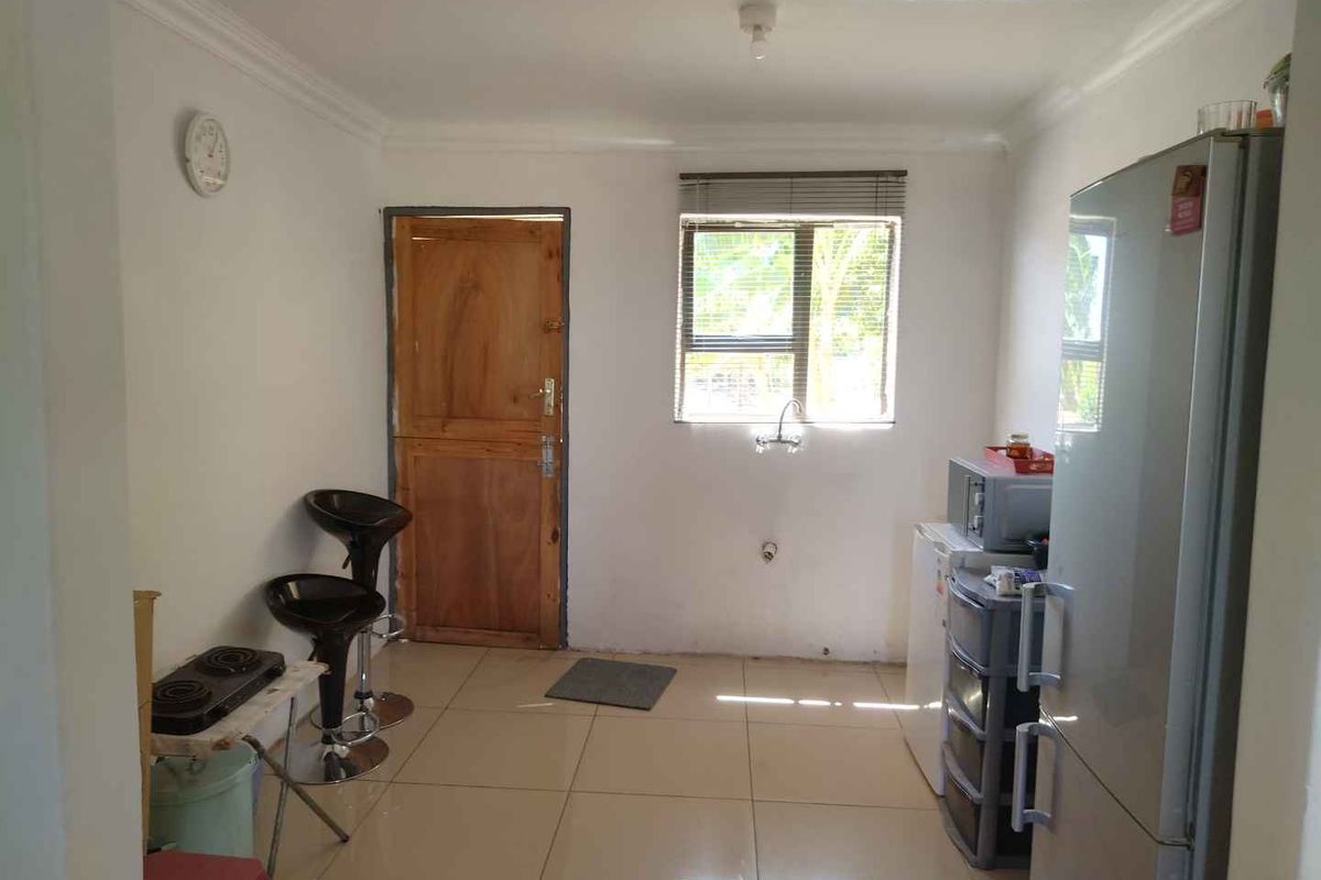 A basic kitchen area featuring tiled flooring, a window with blinds, and a wooden door, currently lacking fitted cabinetry and integrated appliances.