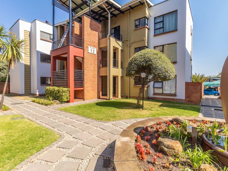 A well-maintained apartment complex exterior featuring modern brick and render facades, landscaped garden areas, and paved walkways leading to the building entrance.