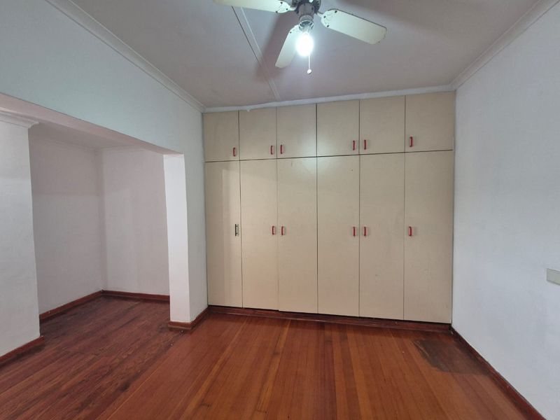 An empty bedroom featuring original hardwood flooring, extensive built-in cupboard storage, and a ceiling fan, offering a blank canvas for renovation.