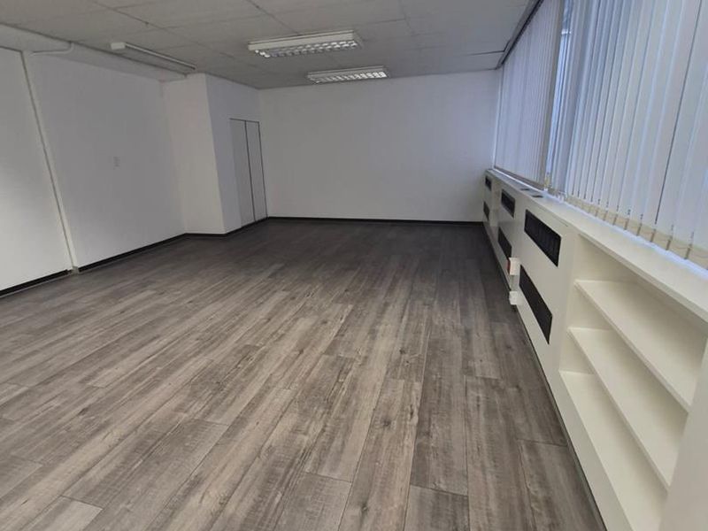 A bright, empty office space featuring modern wood-look laminate flooring, suspended ceiling tiles, and integrated shelving units along the window wall.