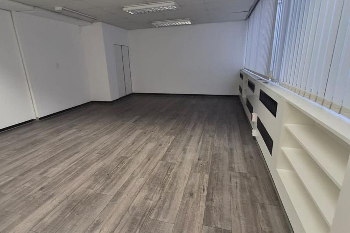 A bright, empty office space featuring modern wood-look laminate flooring, suspended ceiling tiles, and integrated shelving units along the window wall.