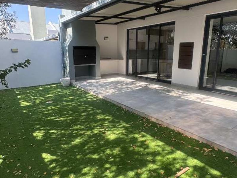 A covered patio area featuring a built-in braai, tiled flooring, and direct access to the garden through sliding doors, shaded by overhead netting.