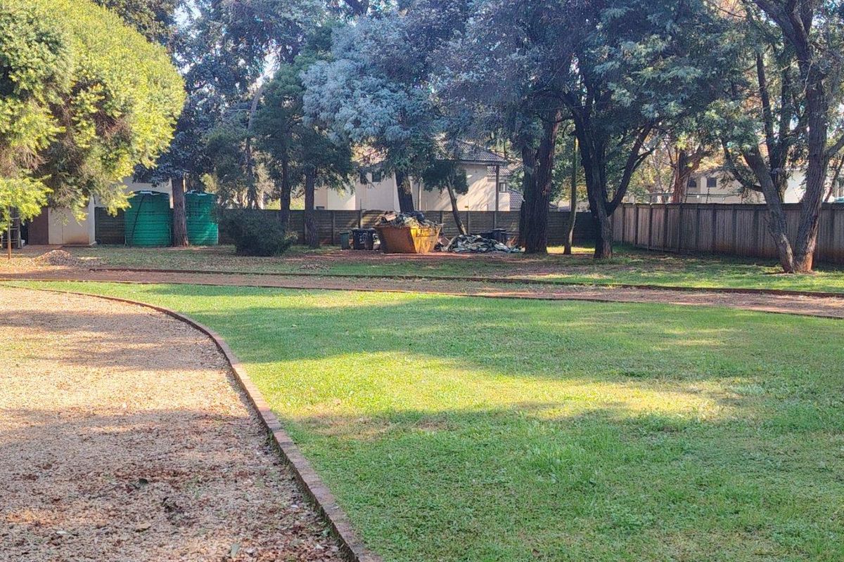 Spacious garden area featuring mature trees, gravel driveway, brick edging, and perimeter timber fencing with installed water storage tanks.