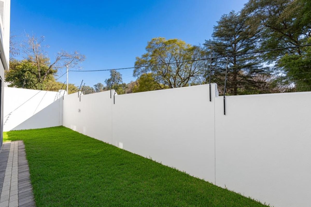 A well-maintained rear garden area featuring a manicured lawn, paved walkway, and high perimeter wall with integrated electric fencing for added security.