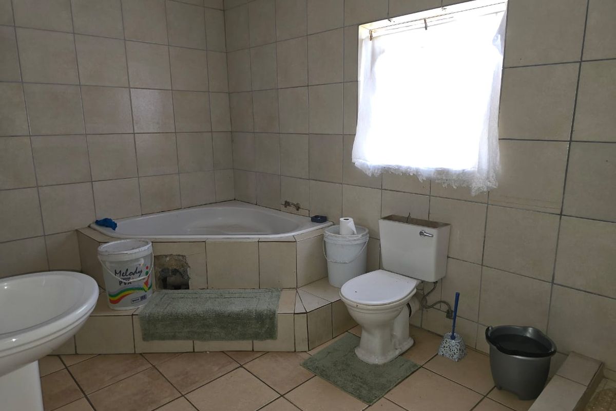 A dated bathroom featuring a corner bathtub, pedestal basin, and toilet, finished with neutral ceramic wall and floor tiles.