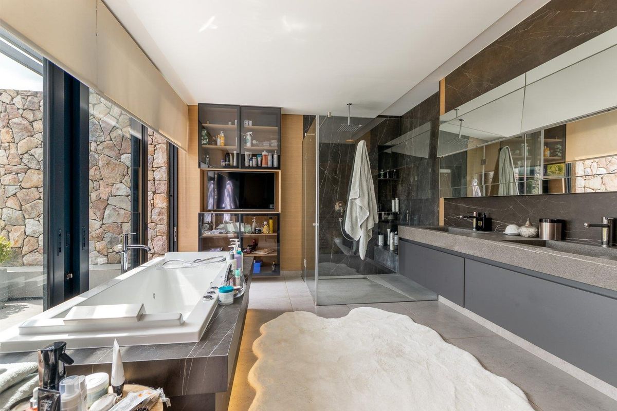 Modern luxury bathroom featuring a freestanding bathtub, walk-in glass shower, dual vanity with stone countertop, and large windows overlooking a stone wall.