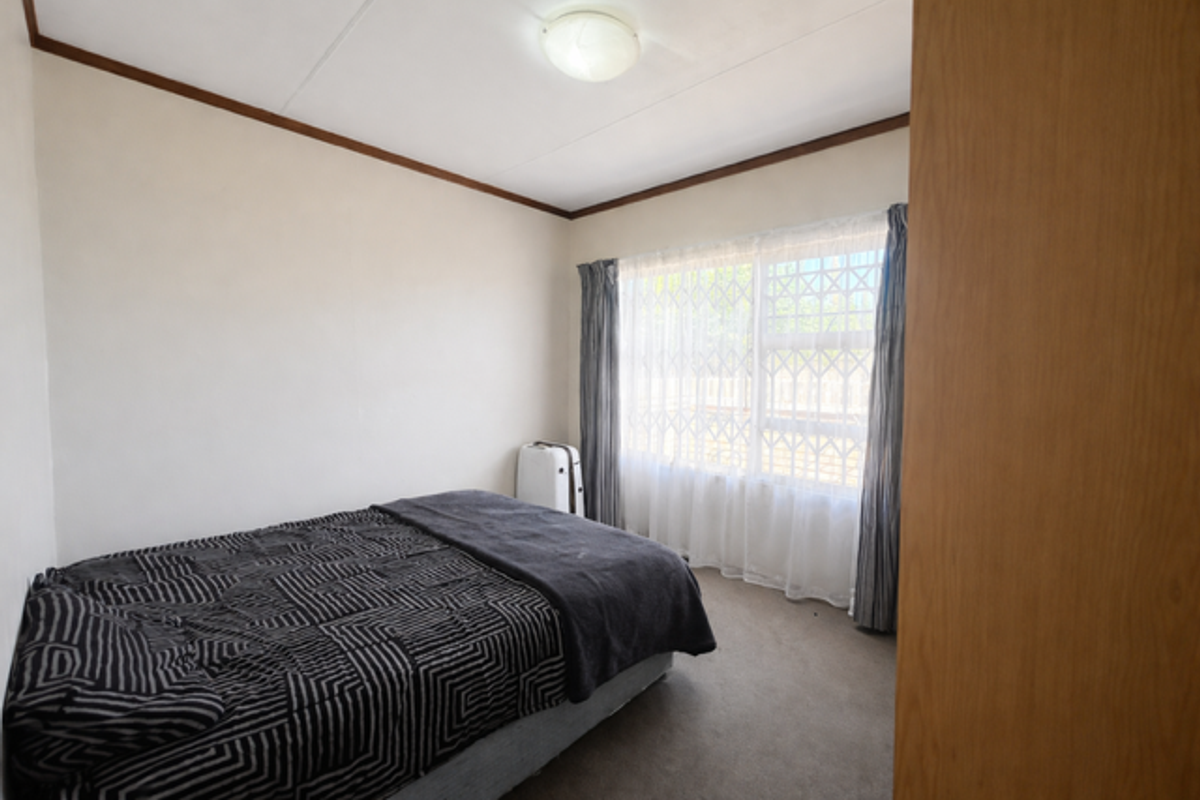 A standard bedroom featuring neutral walls, carpeted flooring, and a window fitted with security bars and curtains.