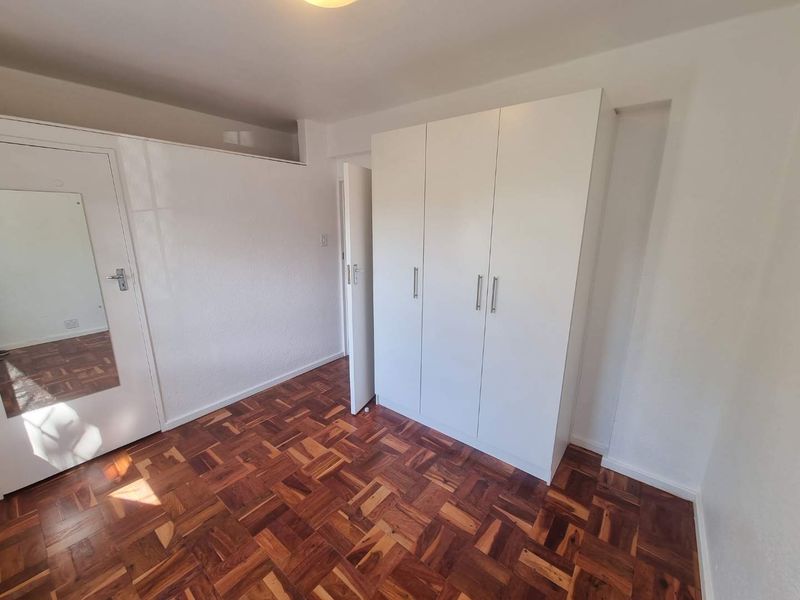 A bright, empty bedroom featuring classic parquet flooring and white built-in cupboards with a mirrored door.