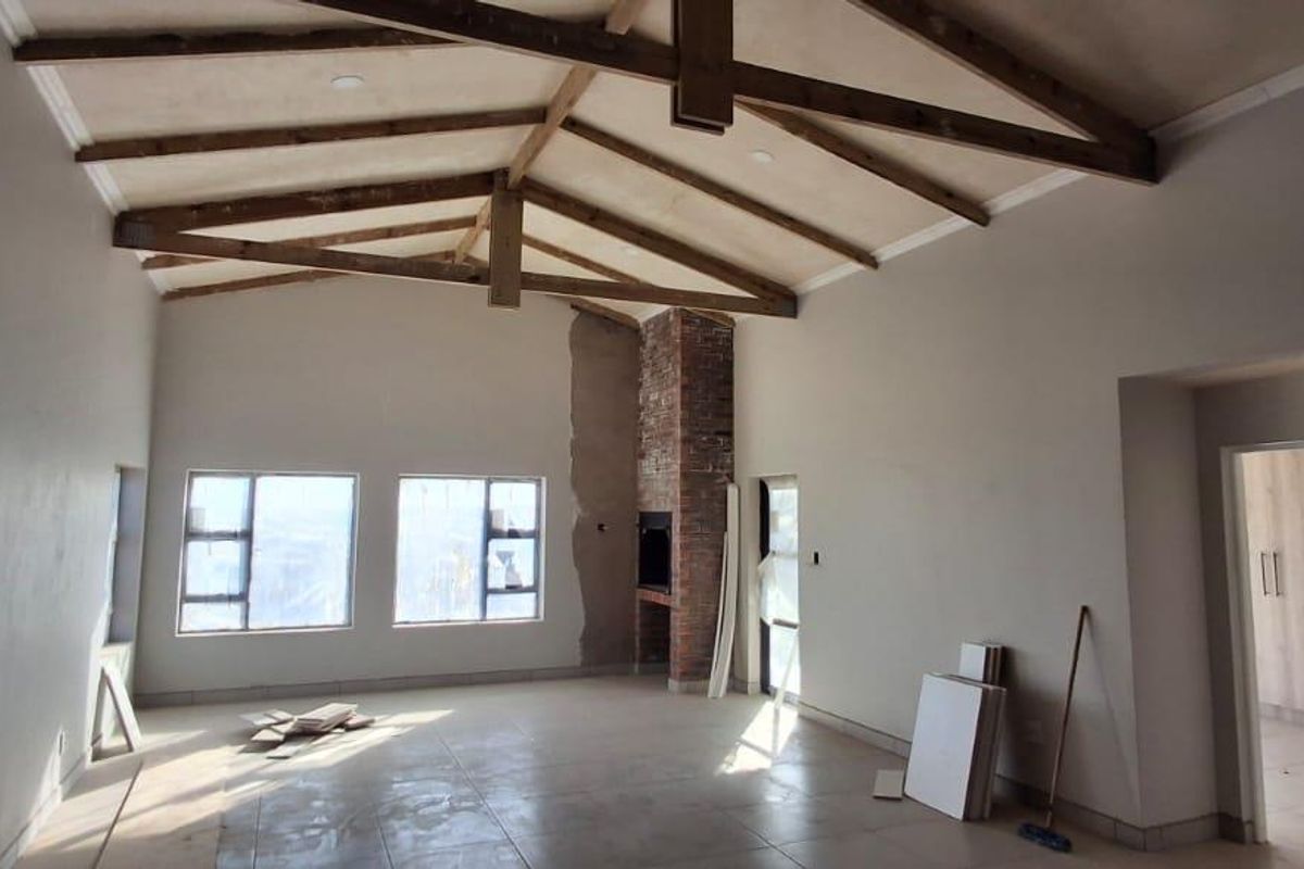 Spacious living area featuring high vaulted ceilings with exposed timber beams, a brick fireplace, and large windows, currently in a final stage of completion.