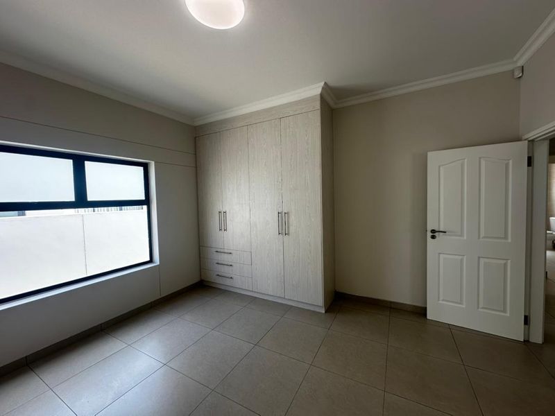 A clean, empty bedroom featuring neutral tiled flooring, built-in cupboards with light wood finish, and a frosted window for privacy.