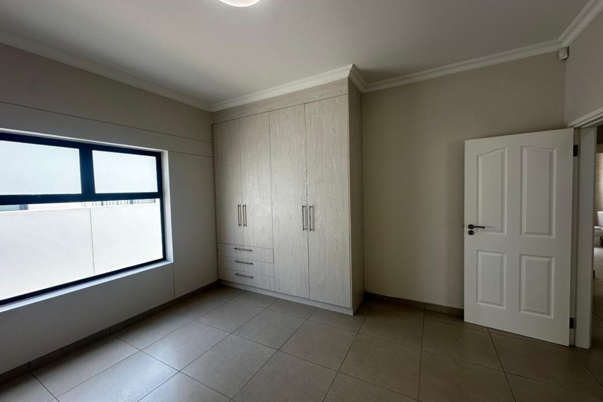 A clean, empty bedroom featuring neutral tiled flooring, built-in cupboards with light wood finish, and a frosted window for privacy.