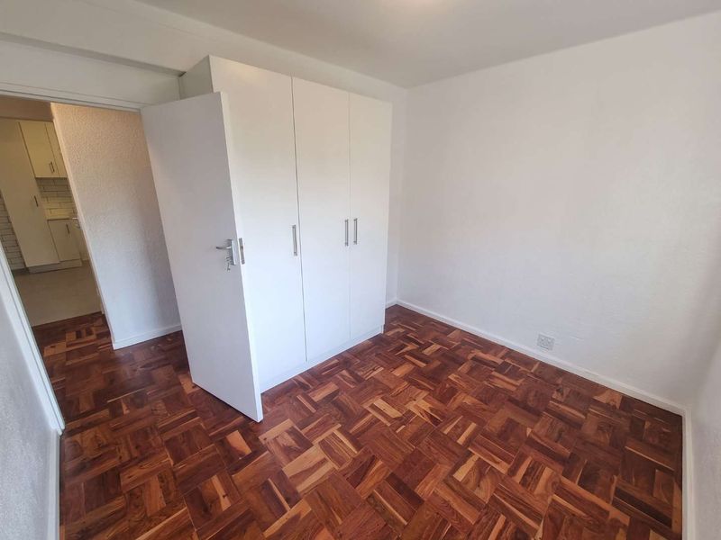 A clean, empty bedroom featuring classic parquet flooring and a white built-in wardrobe, with a view into the adjacent kitchen area.