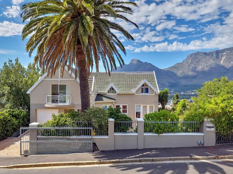 Front elevation of a double-story townhouse featuring a tiled roof, balcony, and secure perimeter wall with a metal gate, set against a mountain backdrop.