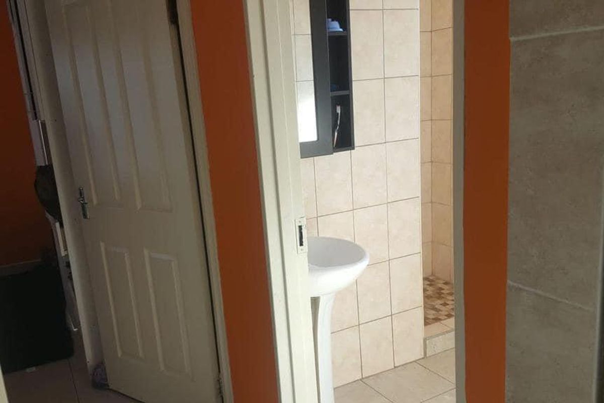 A view into a bathroom featuring a pedestal sink and tiled shower area, accessed via a hallway with orange painted walls.