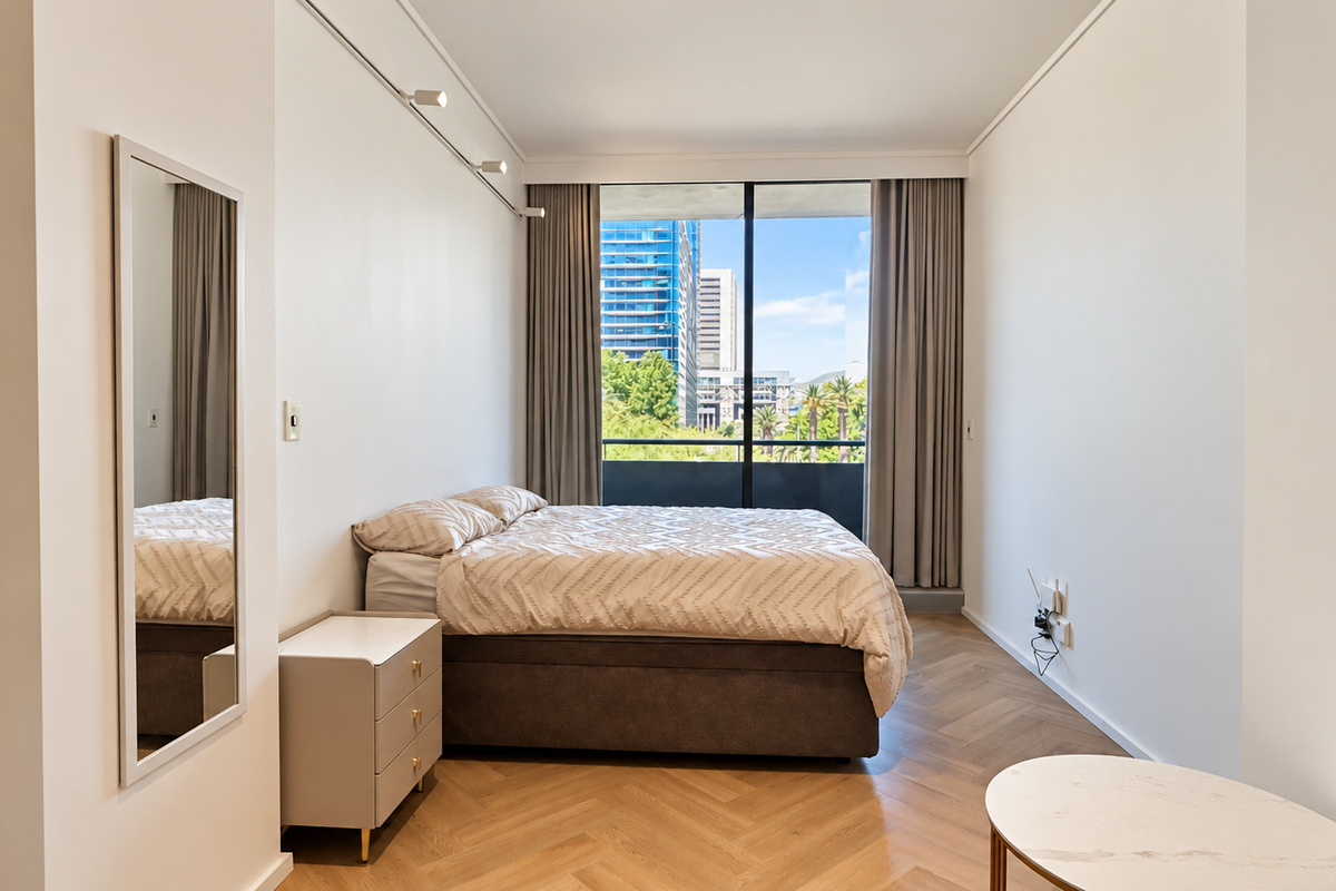 A bright, modern bedroom featuring elegant herringbone timber flooring, track lighting, and a large sliding glass door opening to a balcony with city views.